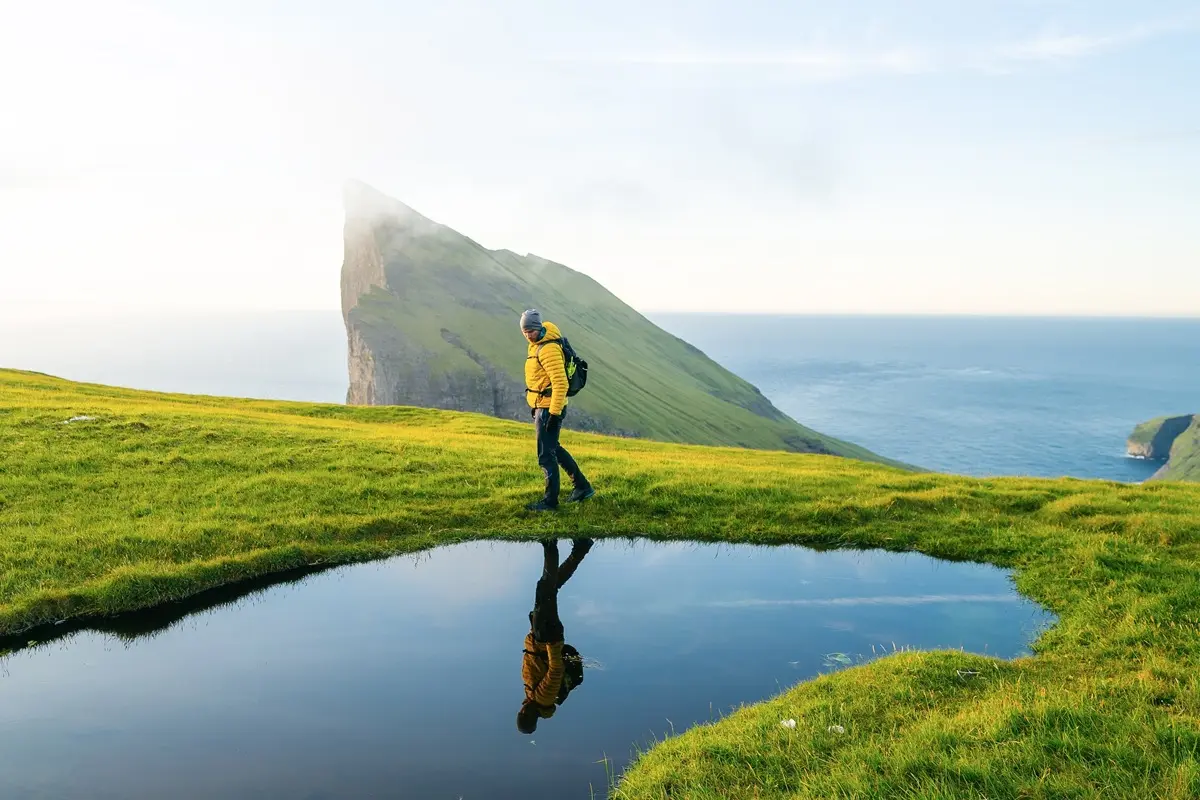 Solo traveler in a yellow jacket with backpack hiking toward Sørvágsvatn on a sunny day