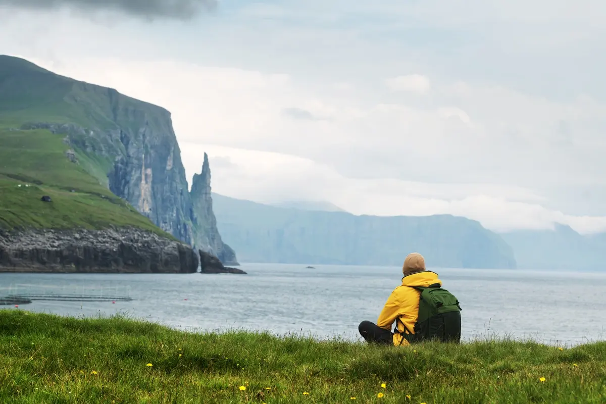 Traveler in yellow jacket with backpack sitting along the Sørvágsvatn hiking trail in the Faroe Islands