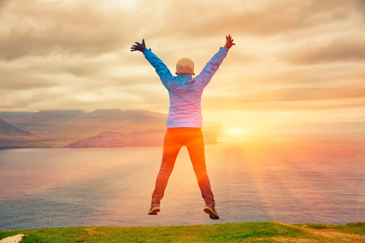 Woman in blue jacket jumping in joy at sunset in the Faroe Islands